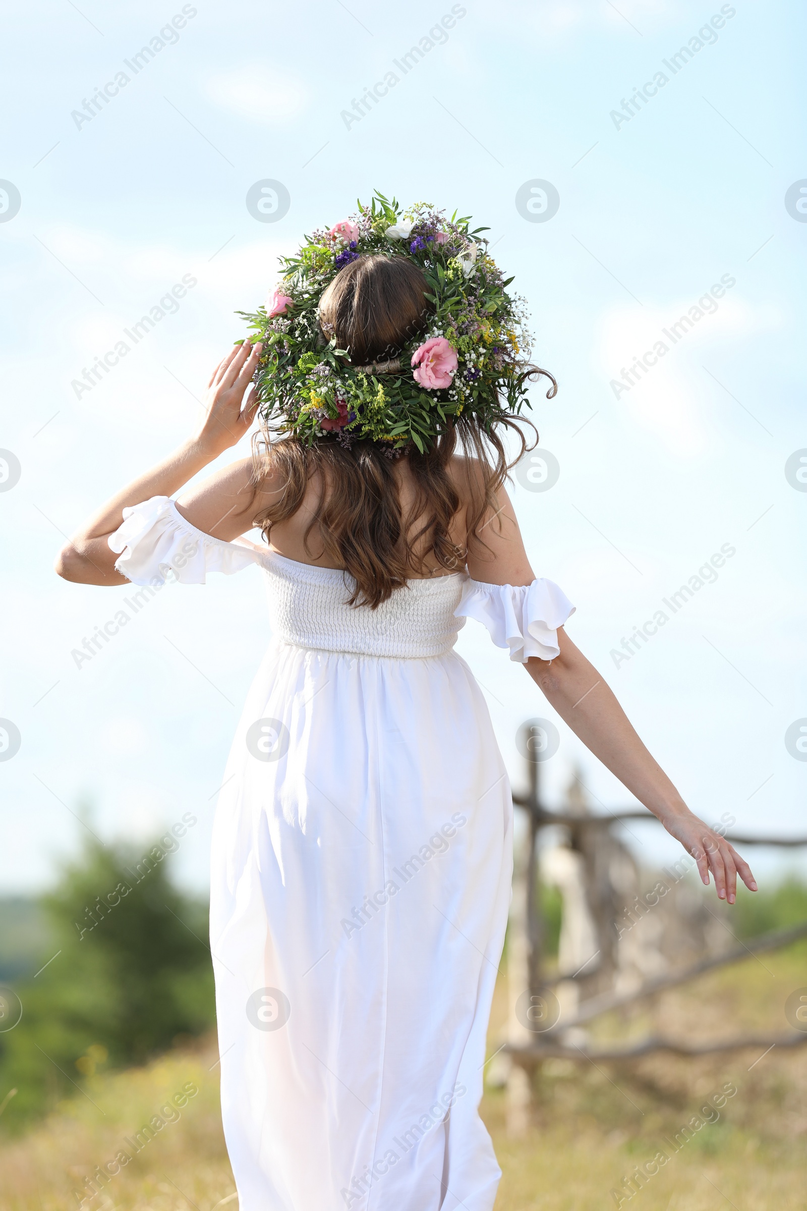 Photo of Young woman wearing wreath made of beautiful flowers outdoors on sunny day, back view