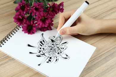 Woman drawing chrysanthemum in sketchbook and flowers at wooden table, closeup