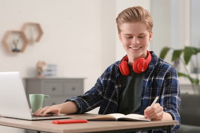 Online learning. Smiling teenage boy writing in notebook near laptop at home