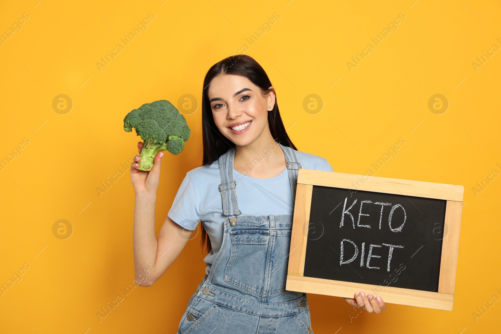 Photo of Happy woman holding broccoli and chalkboard with words Keto Diet on yellow background