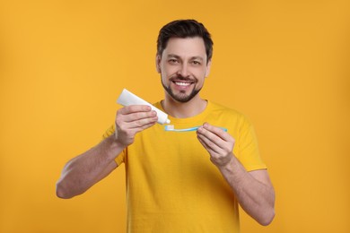 Photo of Happy man squeezing toothpaste from tube onto plastic toothbrush on yellow background