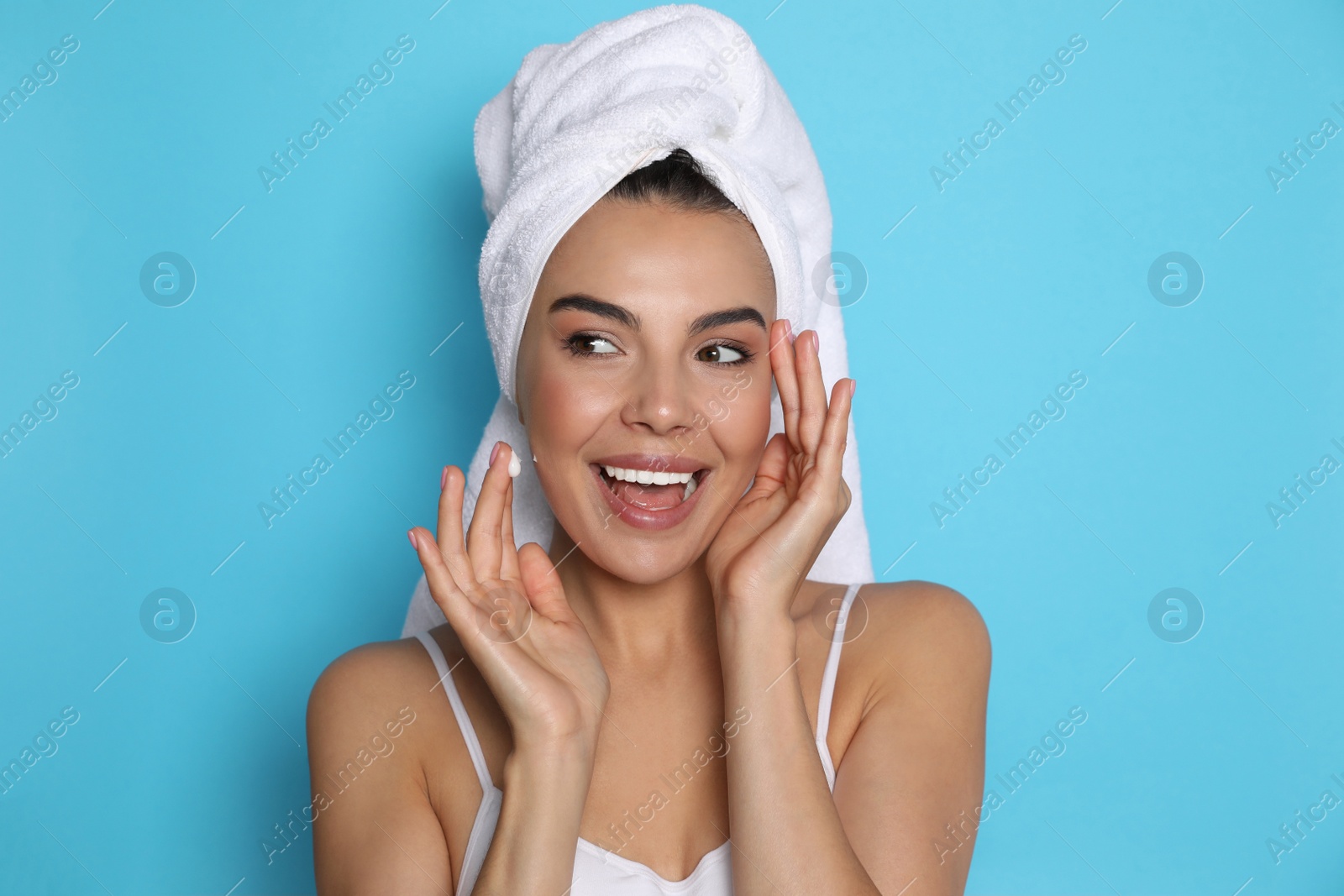 Photo of Beautiful young woman with towel applying cream on face against light blue background