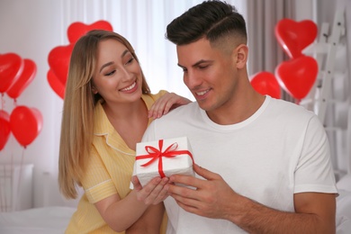 Woman presenting gift to her boyfriend in room decorated with heart shaped balloons. Valentine's day celebration