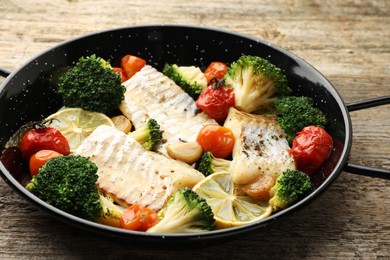 Tasty cod cooked with vegetables in frying pan on wooden table, closeup