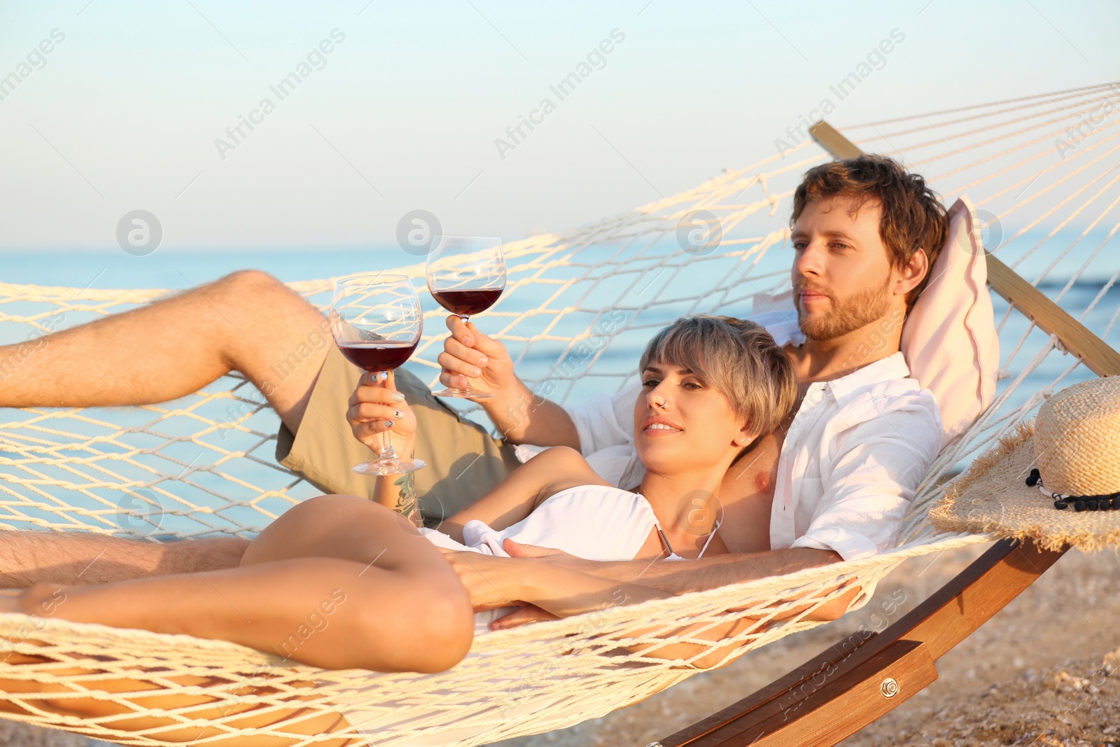 Photo of Young couple resting with glasses of wine in hammock on beach