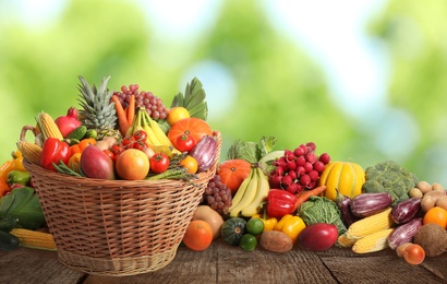 Image of Wicker basket with different fresh organic vegetables and fruits on wooden table against blurred green background