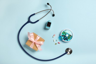 Photo of Stethoscope, pills, box and pulse oximeter on light blue background, flat lay. Medical gift