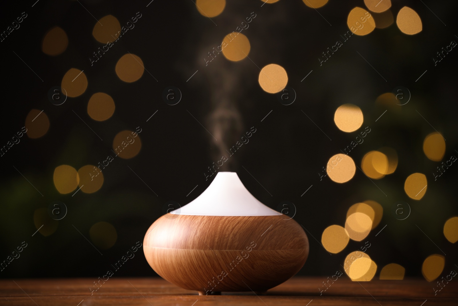 Photo of Modern essential oil diffuser on wooden table against blurred lights, space for text