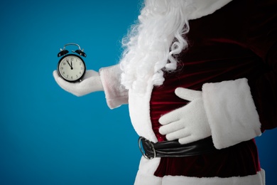 Photo of Santa Claus holding alarm clock on blue background, closeup. Christmas countdown