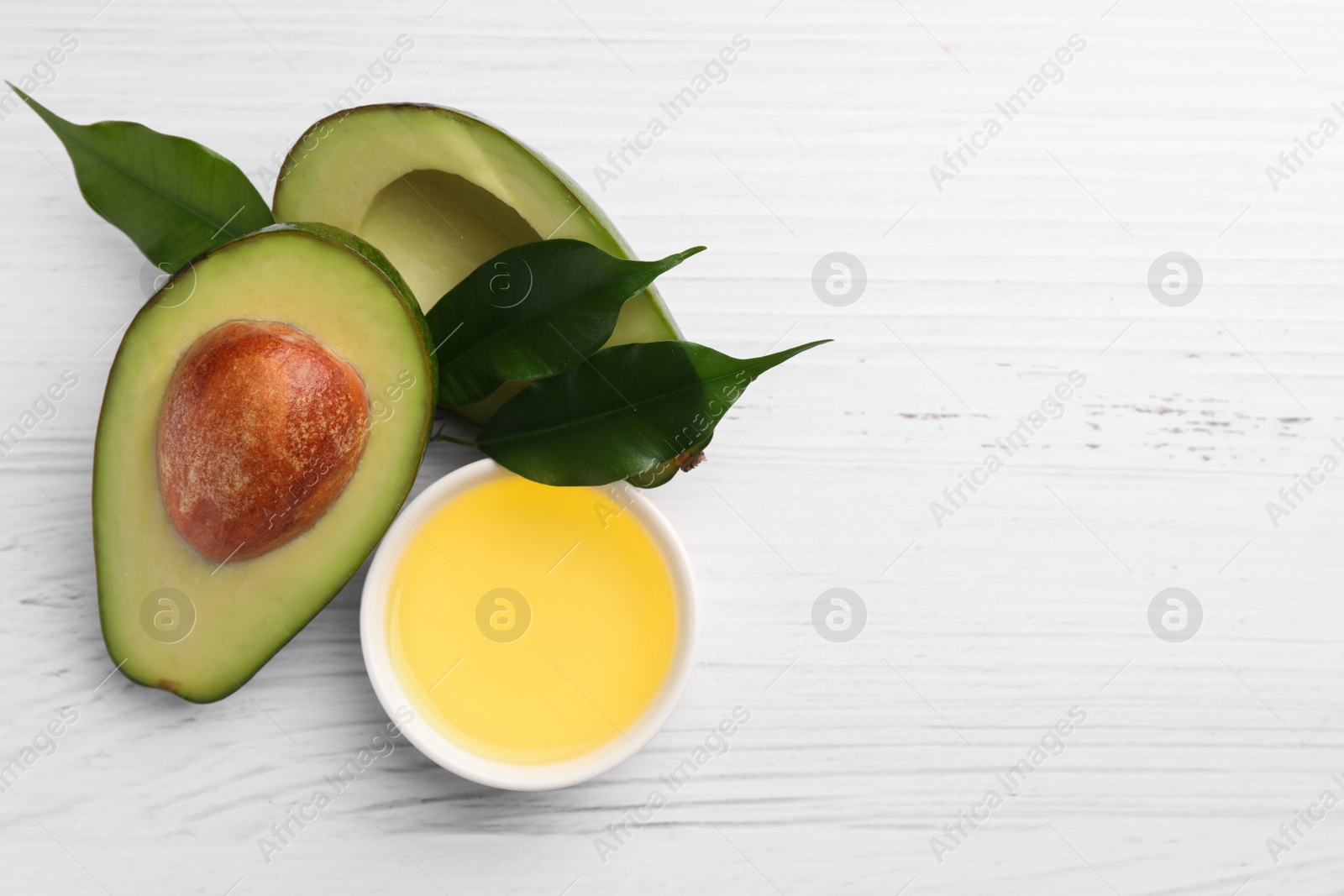 Photo of Cooking oil in bowl and fresh avocados on white wooden table, flat lay. Space for text