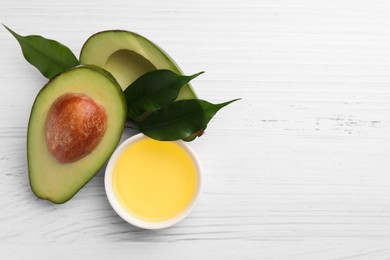 Cooking oil in bowl and fresh avocados on white wooden table, flat lay. Space for text