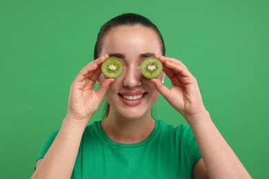 Woman covering eyes with halves of kiwi on green background