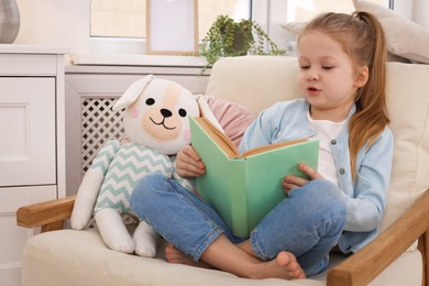 Photo of Cute little girl with toy reading book on armchair at home