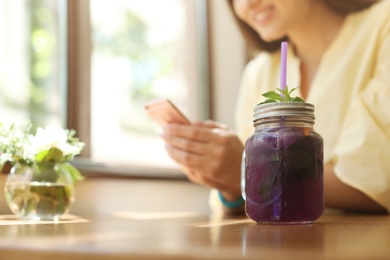 Photo of Mason jar with tasty natural detox lemonade and young woman using mobile phone in cafe