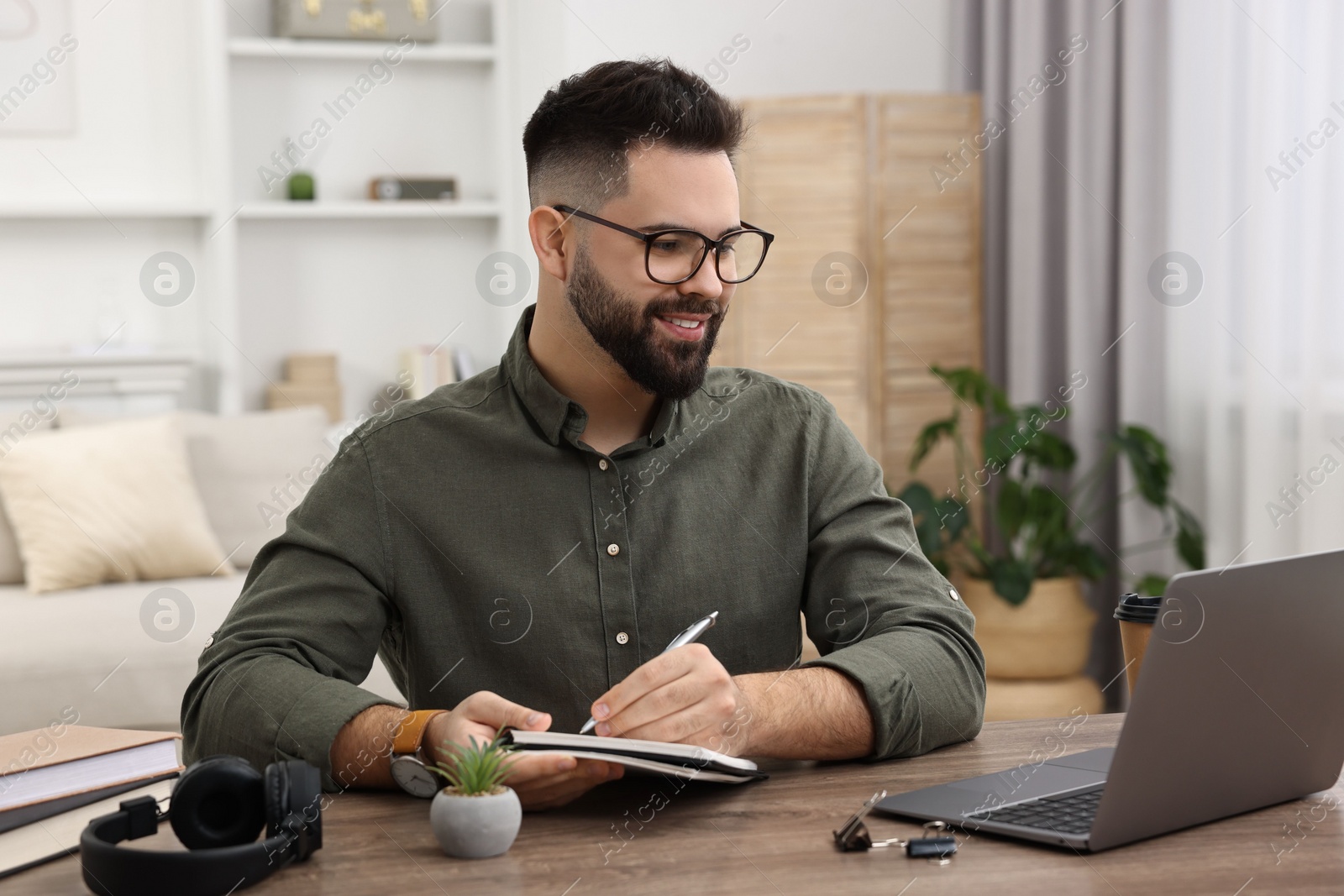 Photo of E-learning. Young man taking notes during online lesson at wooden table indoors