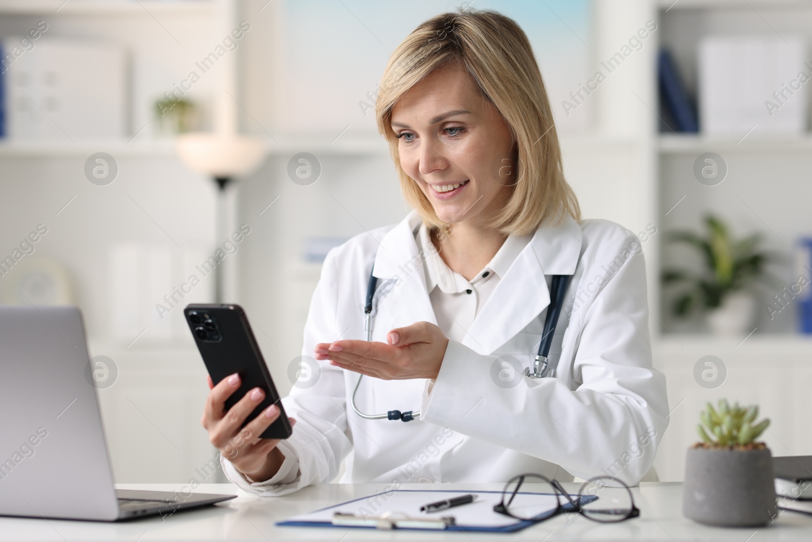 Photo of Smiling doctor with smartphone having online consultation at table in office