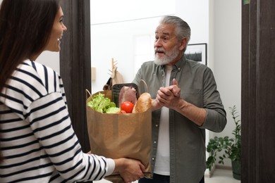 Courier giving paper bag with food products to senior man indoors