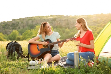 Photo of Young women resting with hot drink and guitar near camping tent in wilderness