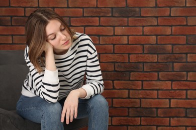 Sad young woman sitting on chair near brick wall, space for text