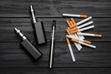 Photo of Cigarettes and different vaping devices on black wooden background, flat lay. Smoking alternative