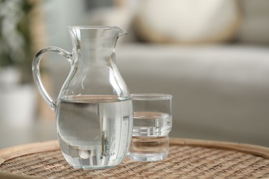 Photo of Jug and glass with clear water on wicker surface against blurred background, closeup