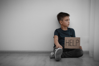 Sad little boy with sign HELP sitting on floor indoors, space for text. Child in danger