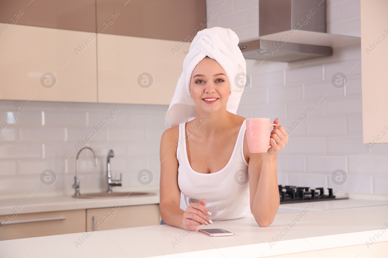 Photo of Beautiful woman with towel on head drinking tea and using smartphone in kitchen