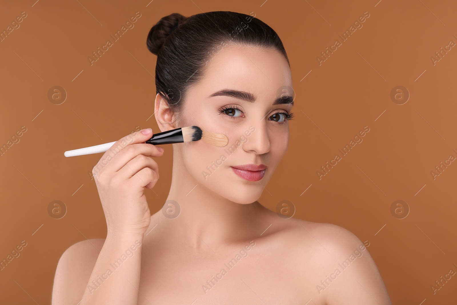 Photo of Woman applying foundation on face with brush against brown background