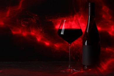Photo of Tasty wine in glass and bottle in red lights on black background, space for text