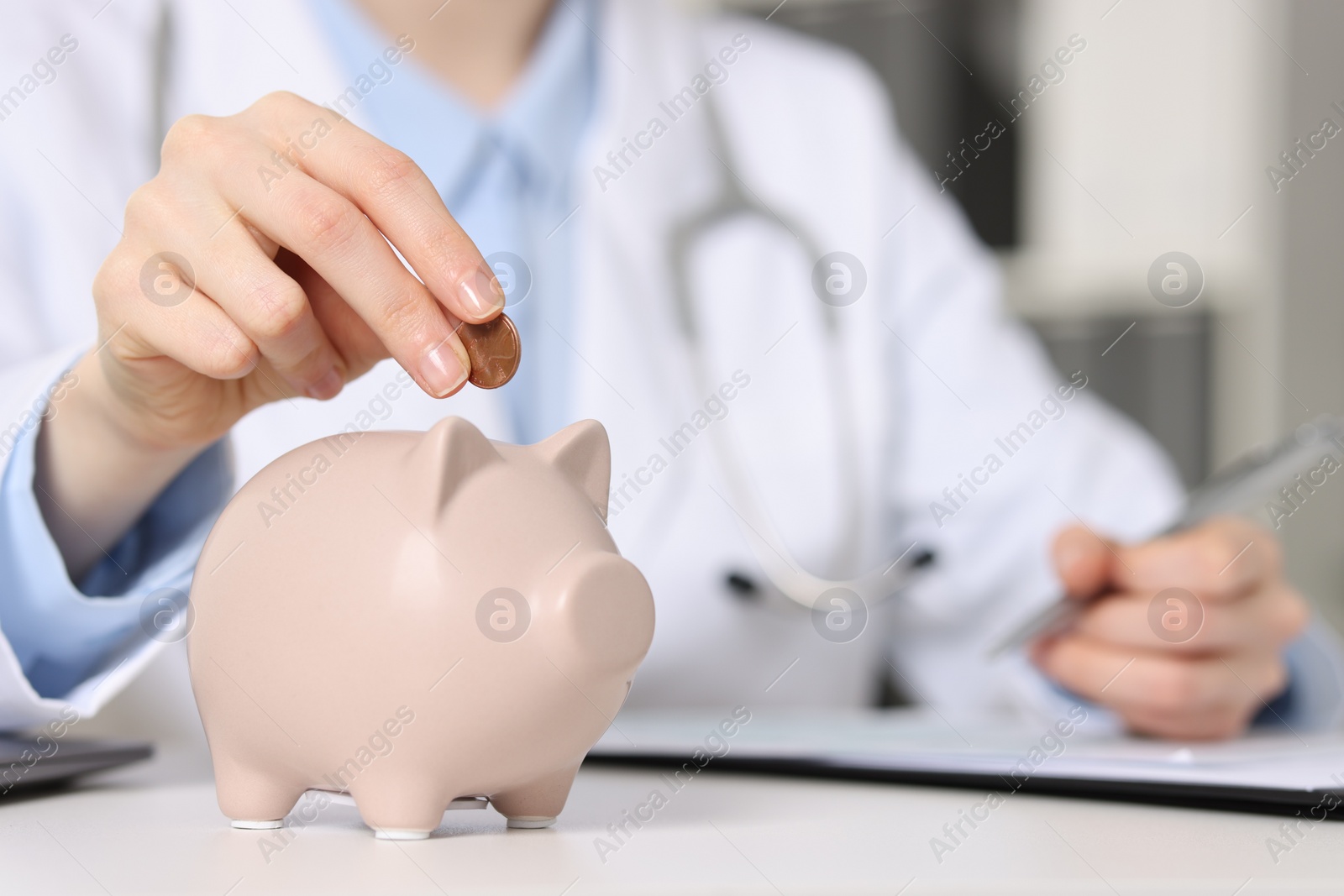 Doctor putting coin into piggy bank while making notes at white table indoors, closeup Photo of Doctor putting coin into piggy bank while making notes at white table indoors, closeup