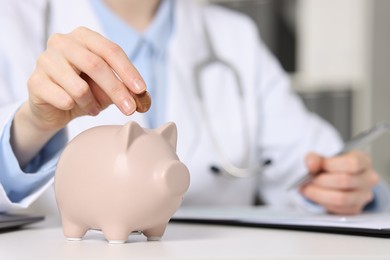 Photo of Doctor putting coin into piggy bank while making notes at white table indoors, closeup