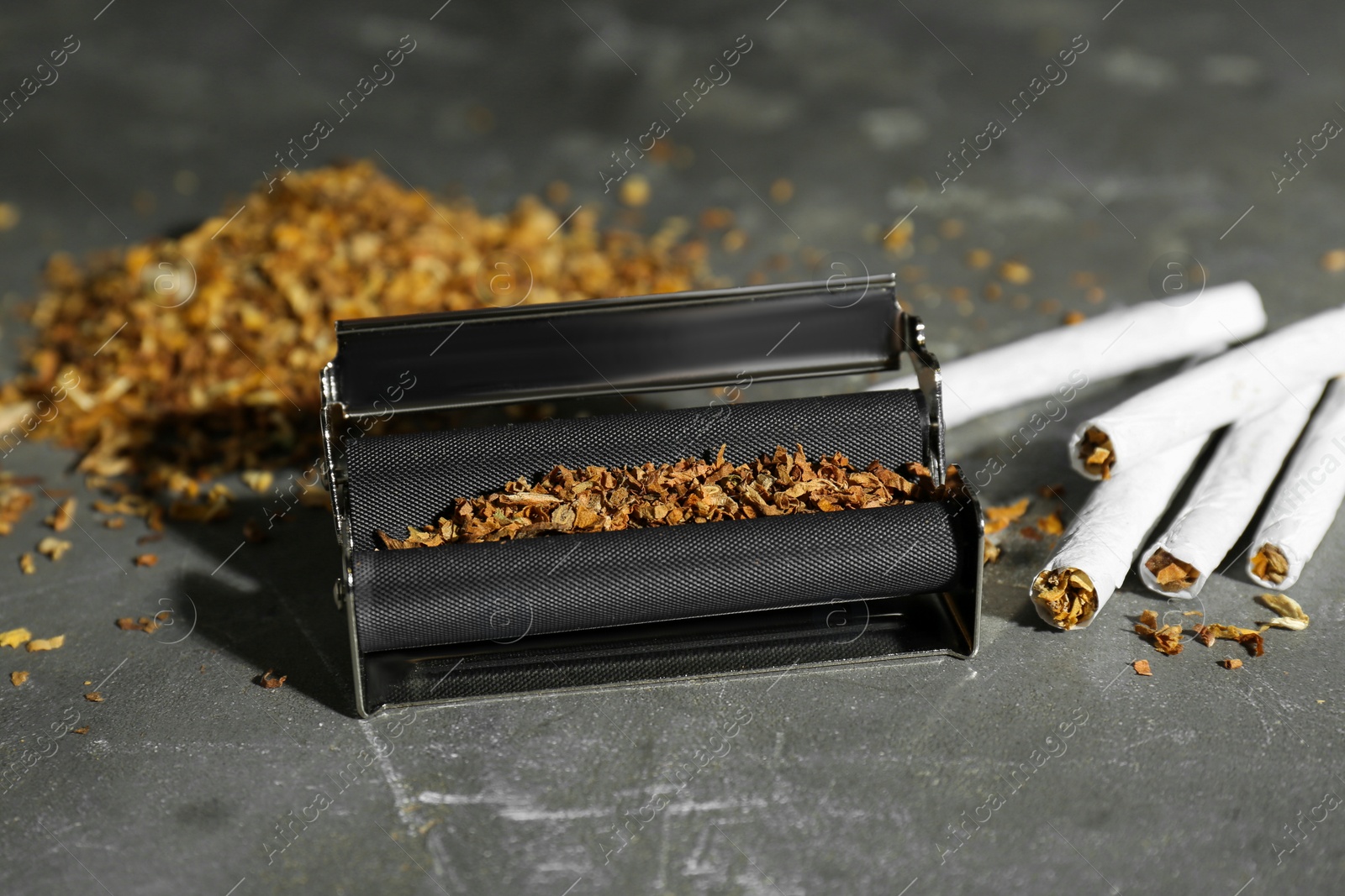 Photo of Roller with tobacco and hand rolled cigarettes on dark grey table