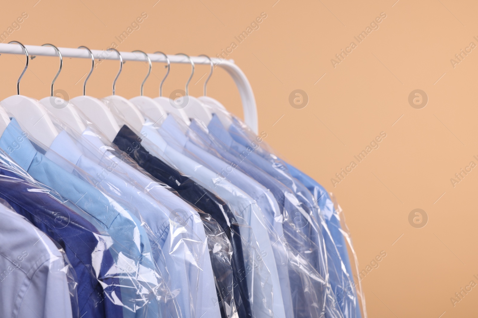 Photo of Dry-cleaning service. Many different clothes in plastic bags hanging on rack against beige background