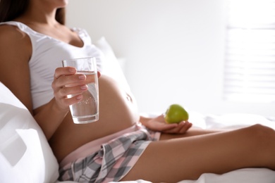 Photo of Young pregnant woman with glass of water and apple in bedroom, closeup. Taking care of baby health