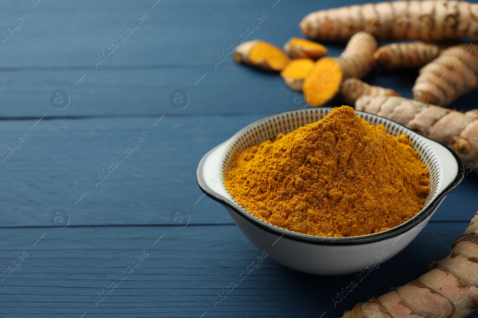 Photo of Bowl with turmeric powder and raw roots on blue wooden table, closeup. Space for text