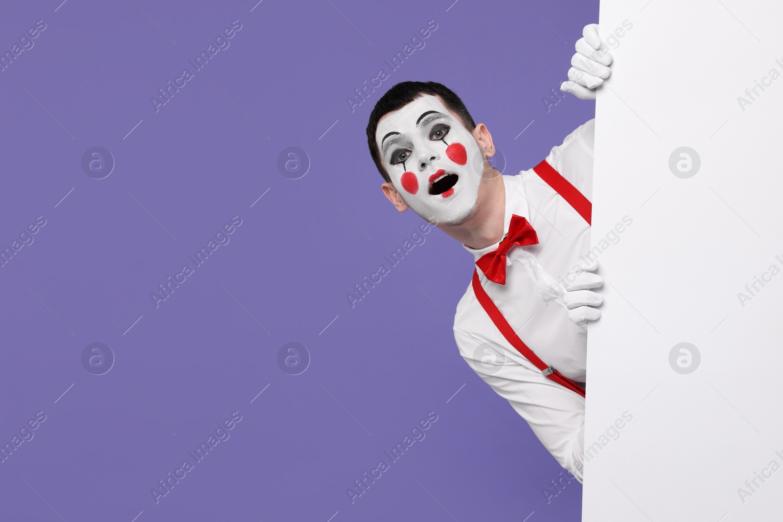 Photo of Funny mime artist peeking out of blank poster on purple background. Space for text