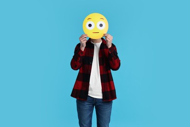 Man covering face with surprised emoticon on light blue background