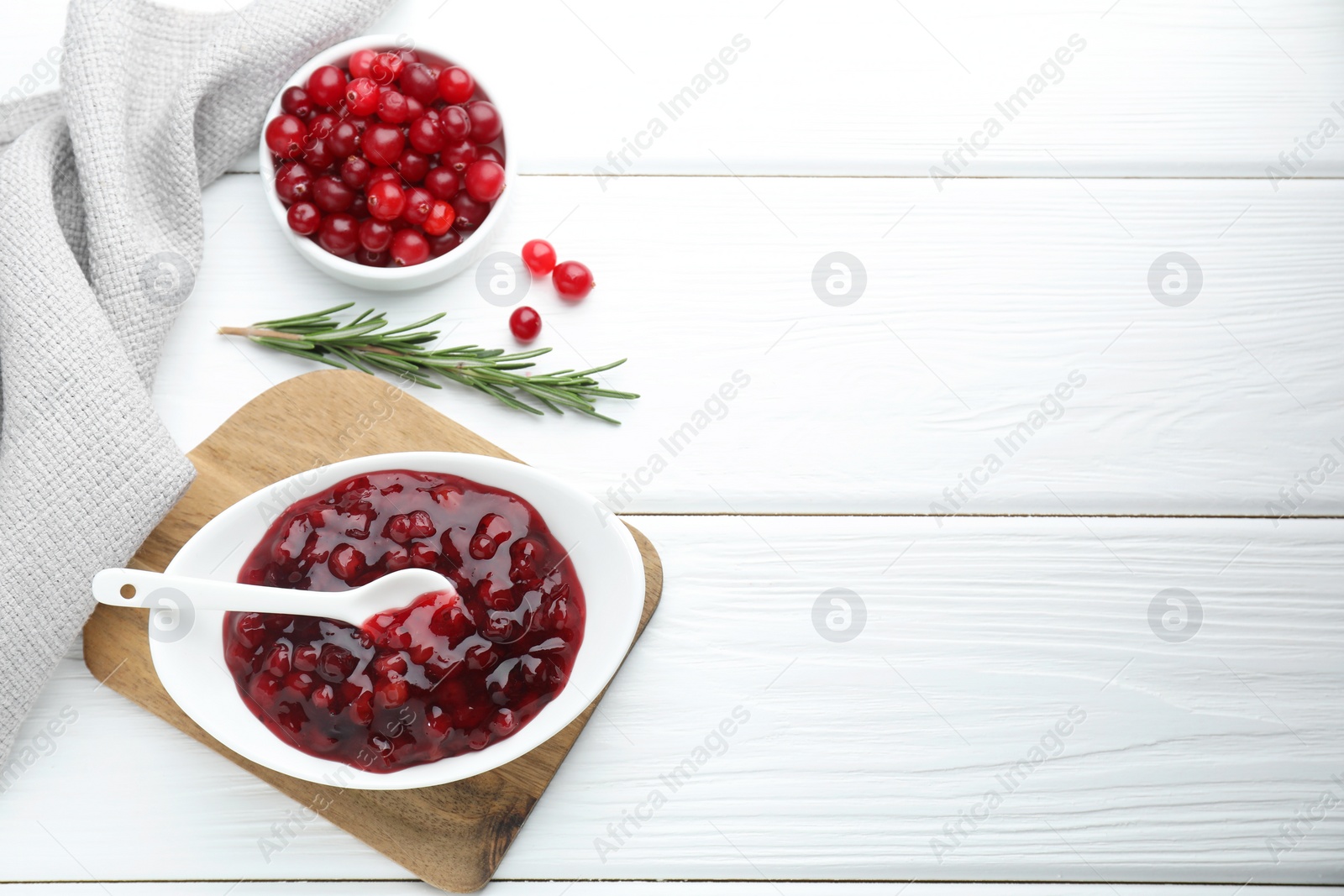 Photo of Tasty cranberry sauce in bowl, fresh berries and rosemary on white wooden table, flat lay. Space for text