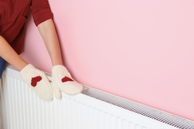 Woman in mittens warming hands on heating radiator near color wall.Space for text