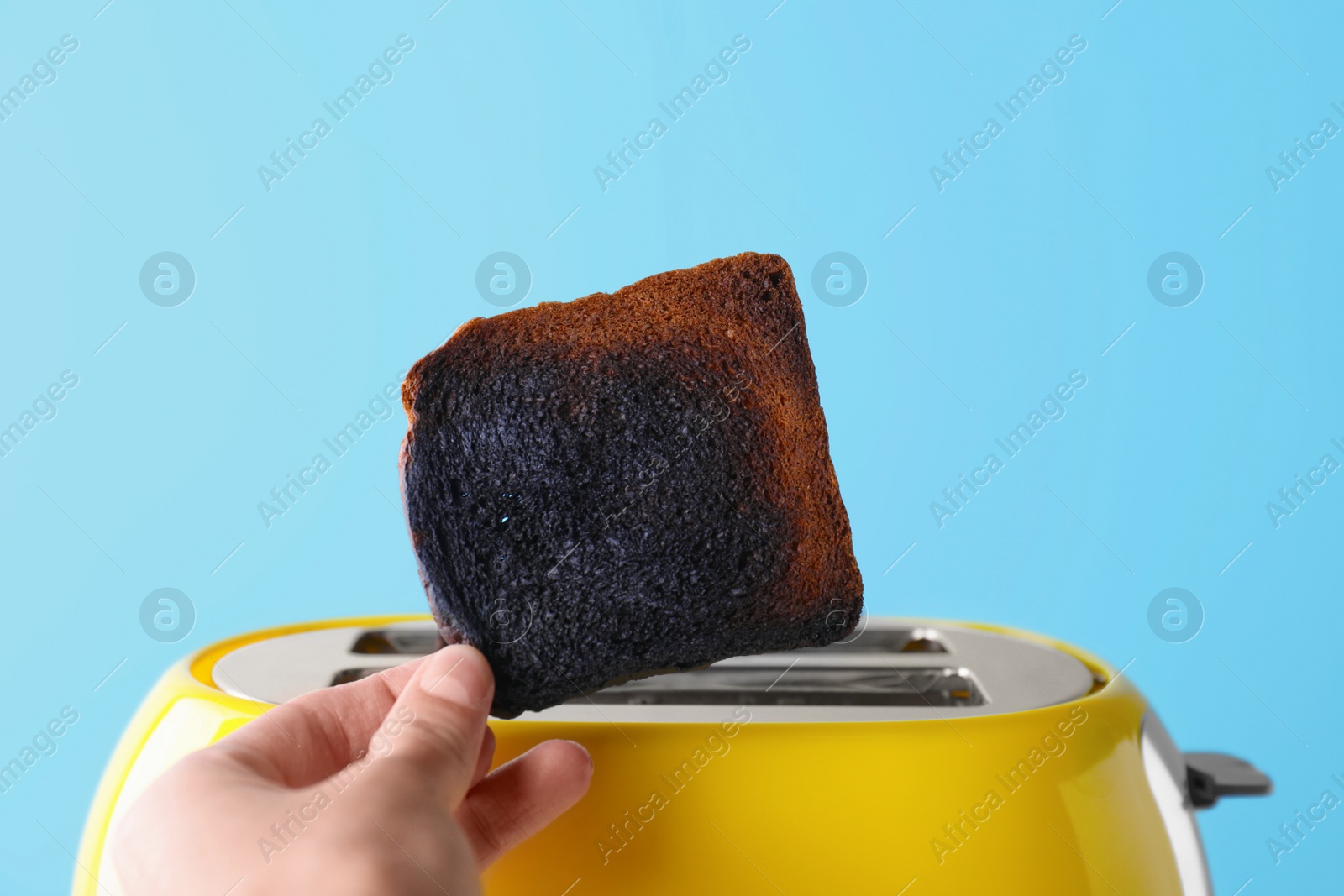 Photo of Woman holding burnt bread near toaster against light blue background, closeup