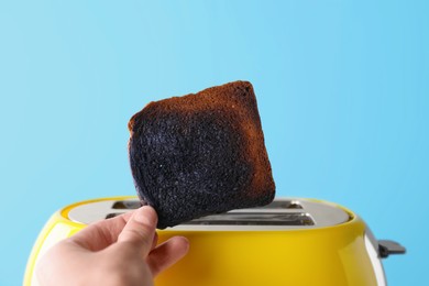Woman holding burnt bread near toaster against light blue background, closeup