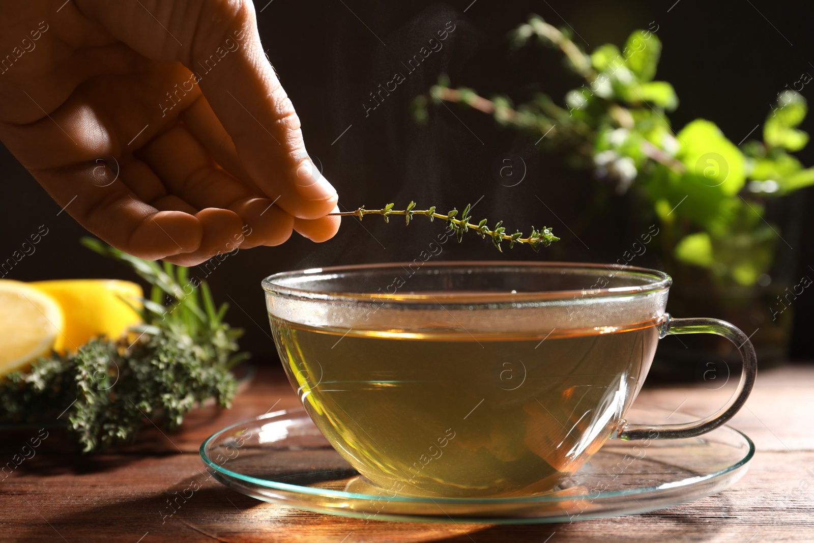 Photo of Woman adding thyme into cup of aromatic herbal tea at wooden table, closeup