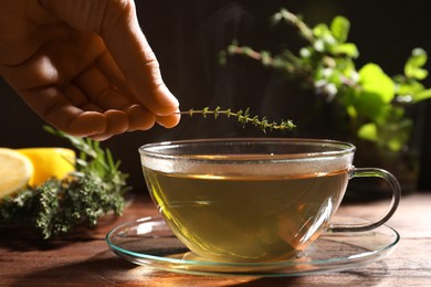 Photo of Woman adding thyme into cup of aromatic herbal tea at wooden table, closeup