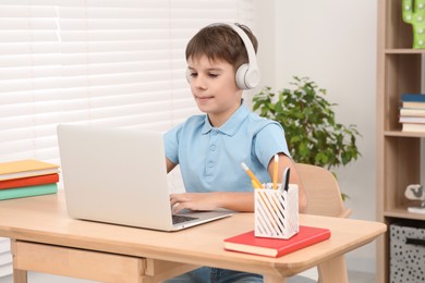 Photo of Boy using laptop and headphones at desk in room. Home workplace