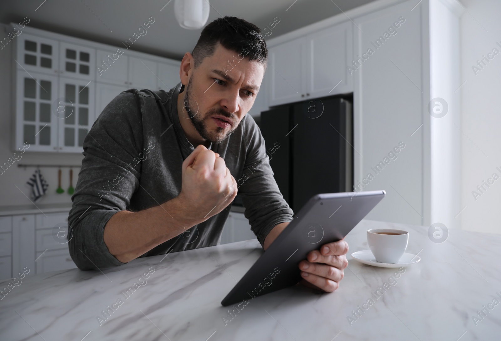 Photo of Emotional man with tablet at table in kitchen. Online hate concept