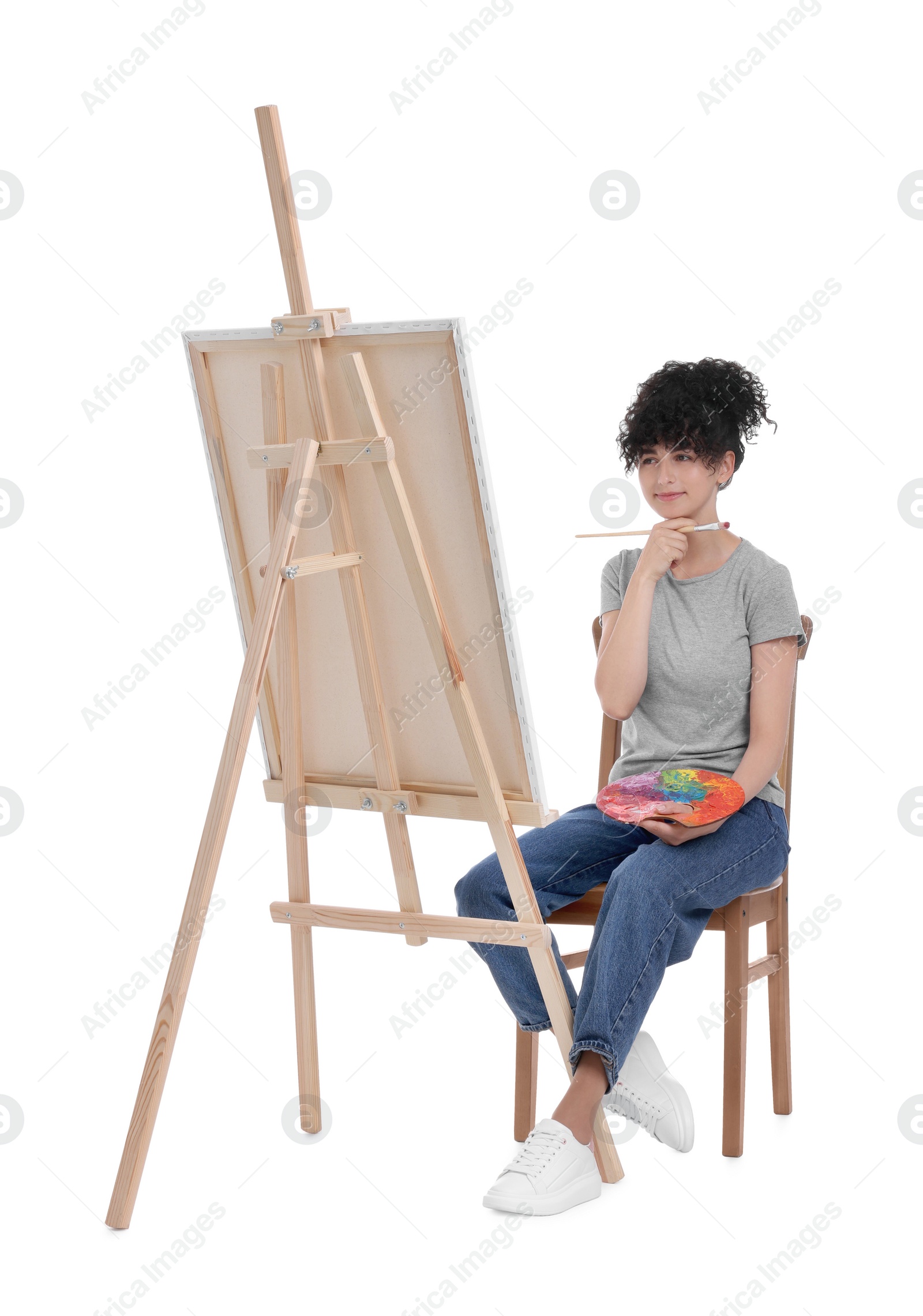 Photo of Young woman holding brush and artist`s palette near easel with canvas against white background