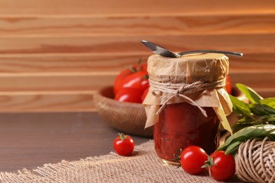 Photo of Jar of tasty tomato paste with spoon, ingredients and thread on wooden table. Space for text