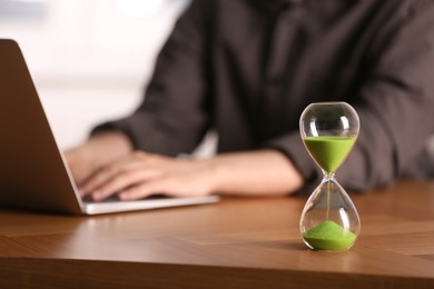 Hourglass with flowing sand on wooden table, selective focus. Man using laptop indoors