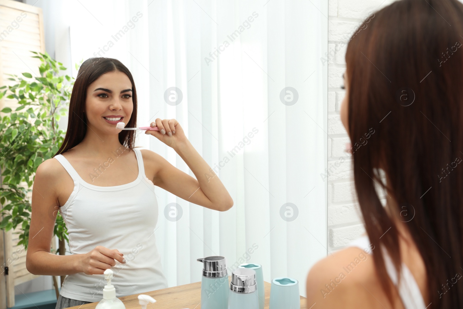 Photo of Beautiful woman brushing teeth near mirror in bathroom at home