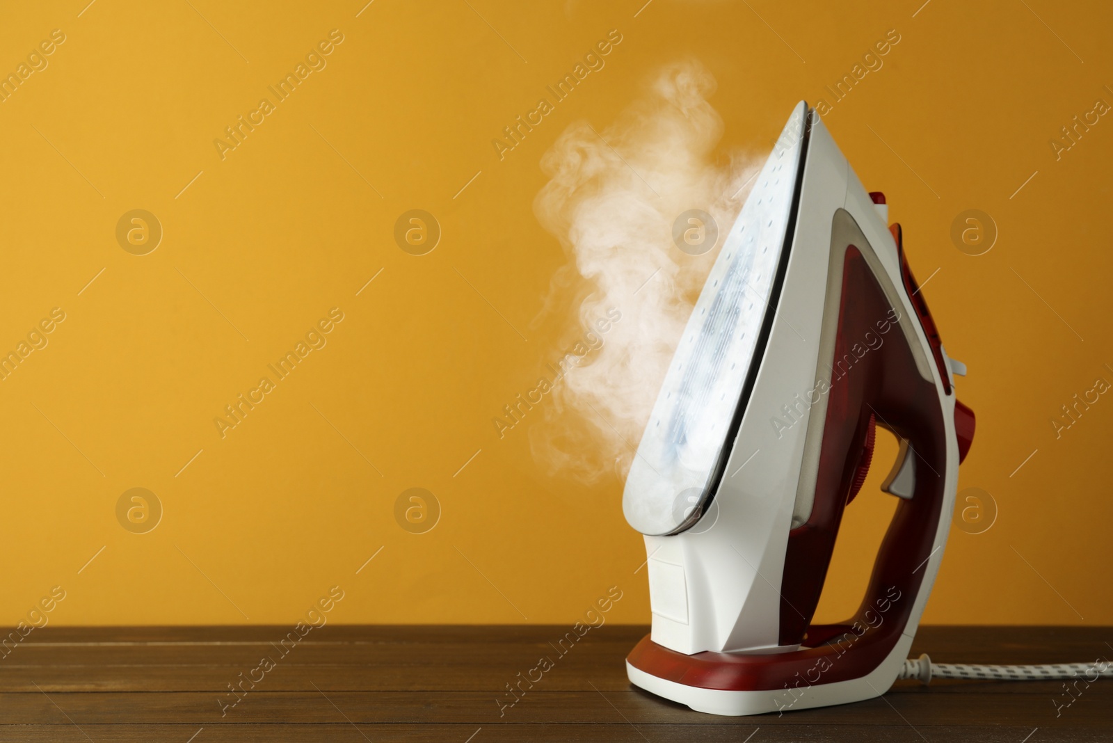 Image of Modern iron with steam on wooden table against orange background, space for text